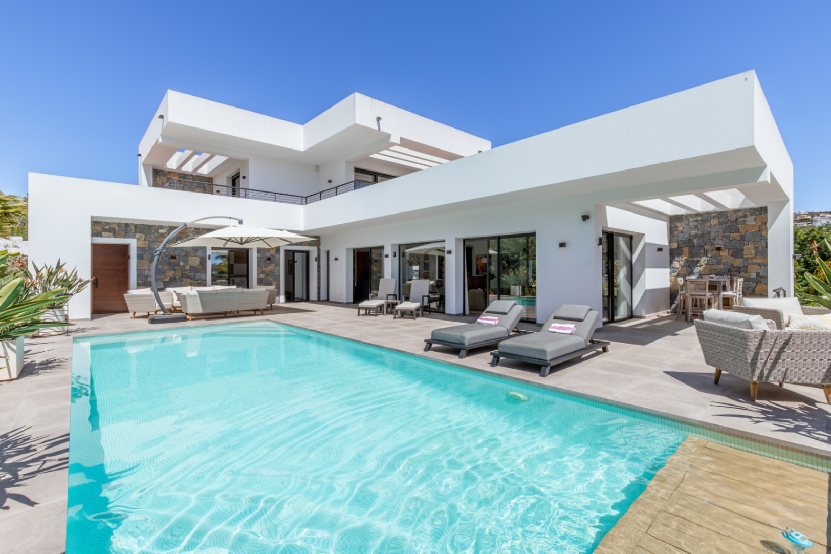 Außenansicht der Villa mit großem Pool, weitläufiger Terrasse und moderner weißer Architektur, kombiniert mit markanten Natursteinwänden.
