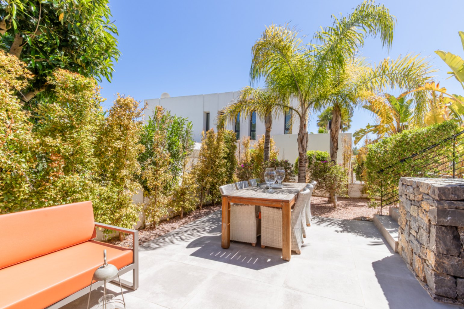 Außenbereich mit Holzesstisch, Korbstühlen und orangefarbenem Sofa, eingebettet in einen Garten mit Palmen und dichter mediterraner Vegetation.