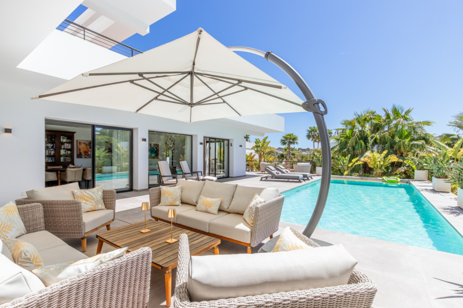 Außenbereich mit Loungemöbeln aus Geflecht und großem Sonnenschirm am Pool, mit direktem Zugang zum Wohnbereich durch bodentiefe Glasschiebetüren.