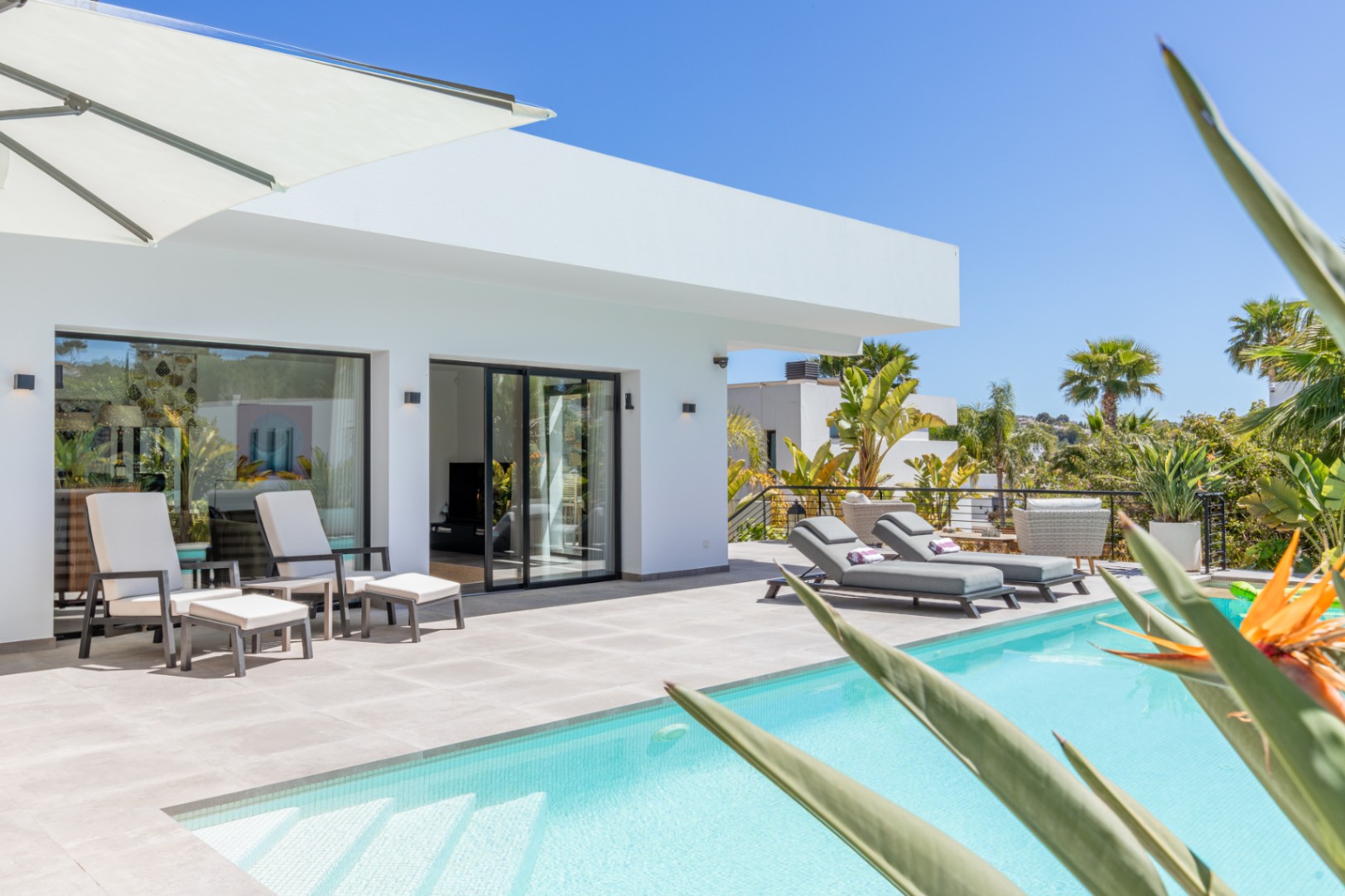 Außenbereich mit Pool, modernen Sonnenliegen und direktem Zugang zum Wohnzimmer durch schwarz gerahmte Glasschiebetüren in minimalistischer weißer Architektur.