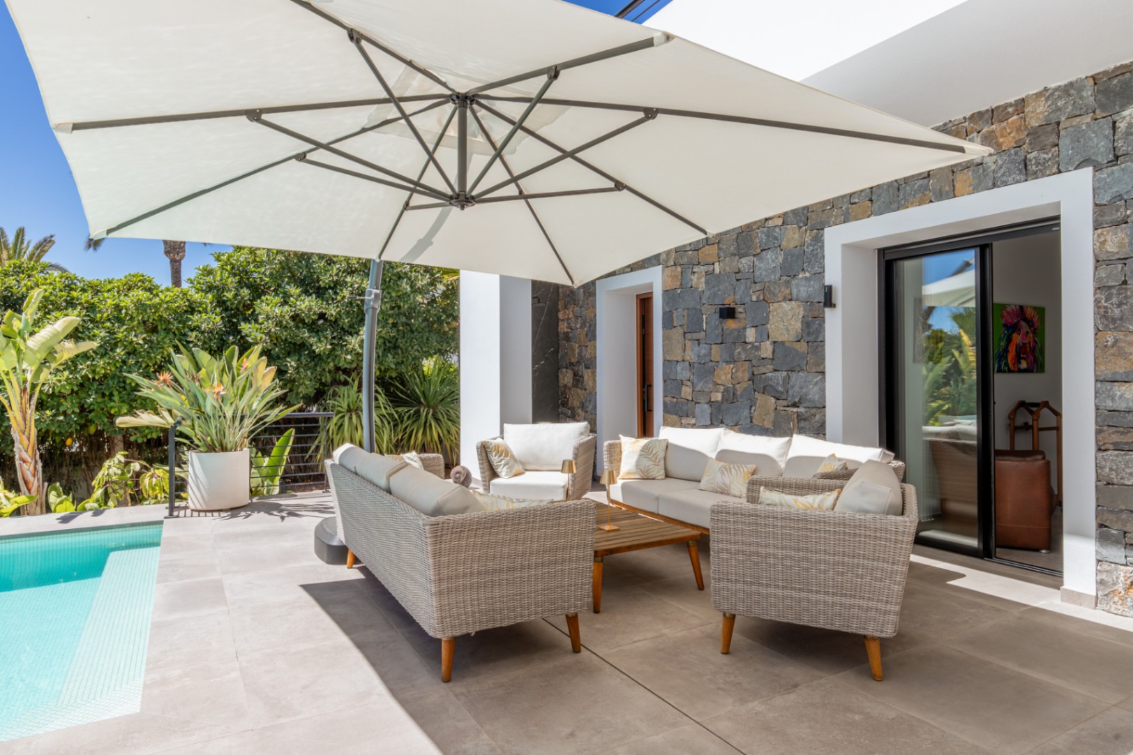 Außenlounge mit Korbmöbeln und großem Sonnenschirm auf einer Fliesenterrasse am Pool, ergänzt durch eine markante Natursteinfassade.