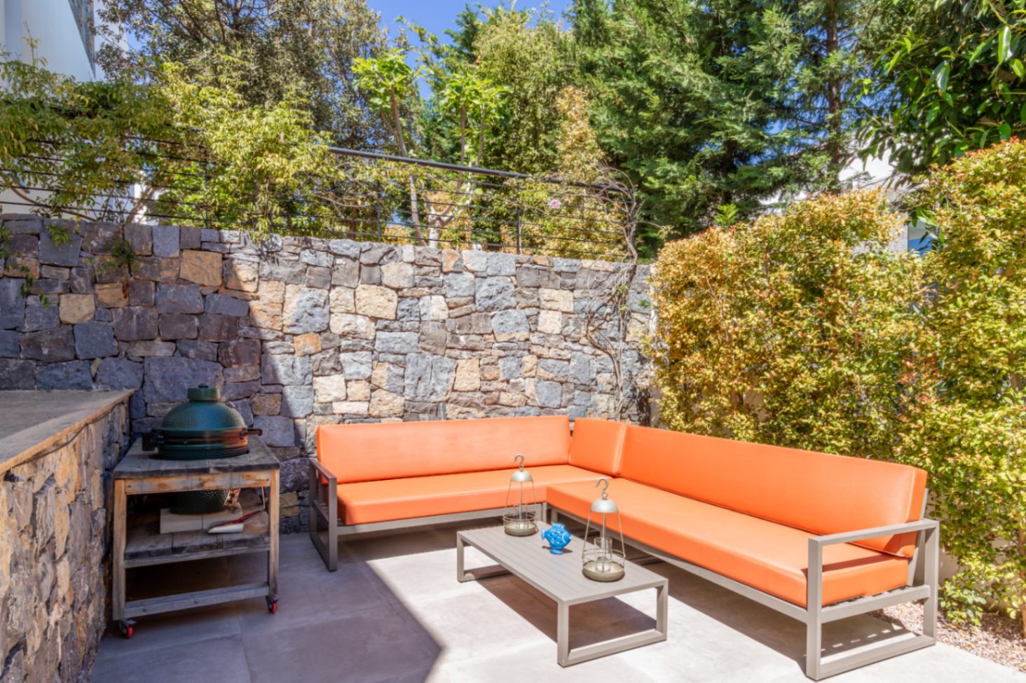Außenlounge mit orangefarbenem Ecksofa, Keramikgrill und Natursteinmauer, umgeben von privatem, mediterranem Bewuchs.