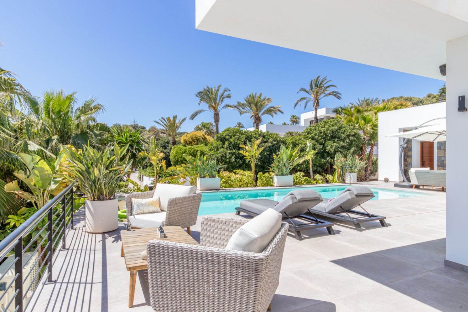 Außenterrasse mit Rattanmöbeln und Sonnenliegen am Pool, eingebettet in einen mediterranen Garten mit Palmen und dichter Bepflanzung.