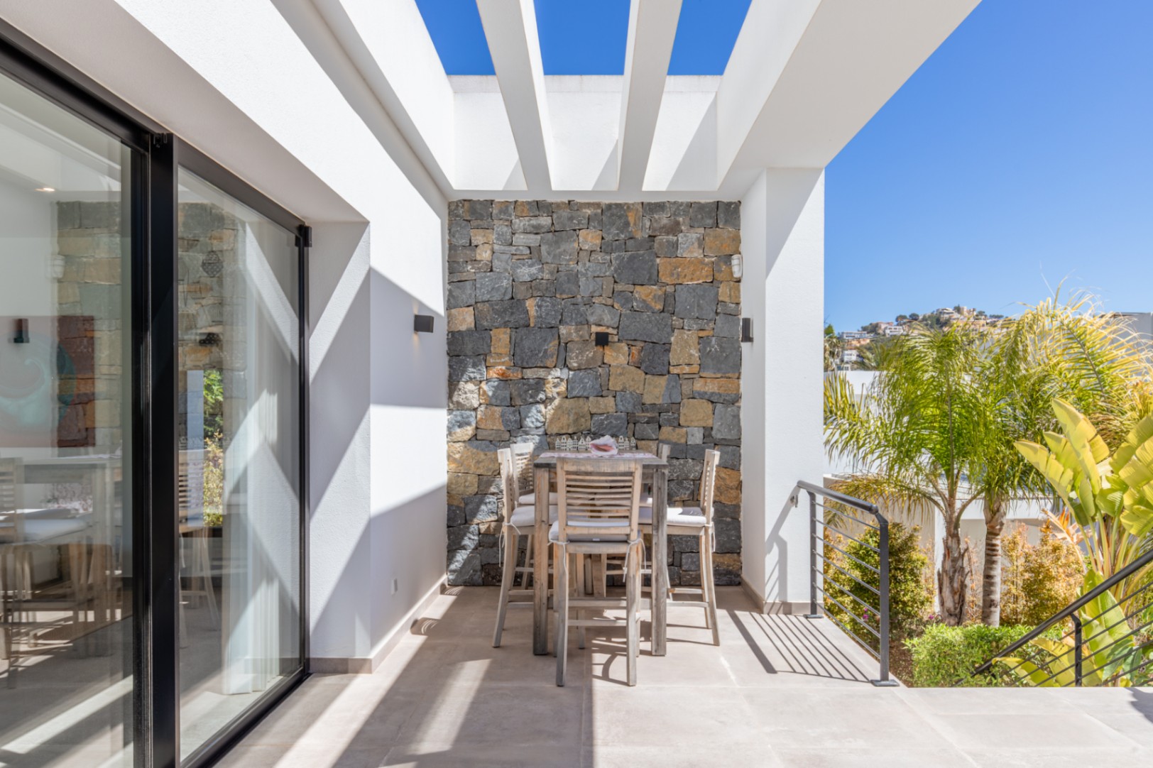 Außenunterrasse mit weißer Pergola, Natursteinwand und hohem Esstisch aus Holz. Große Glasschiebetüren verbinden den hellen Außenbereich mit dem Wohnbereich.