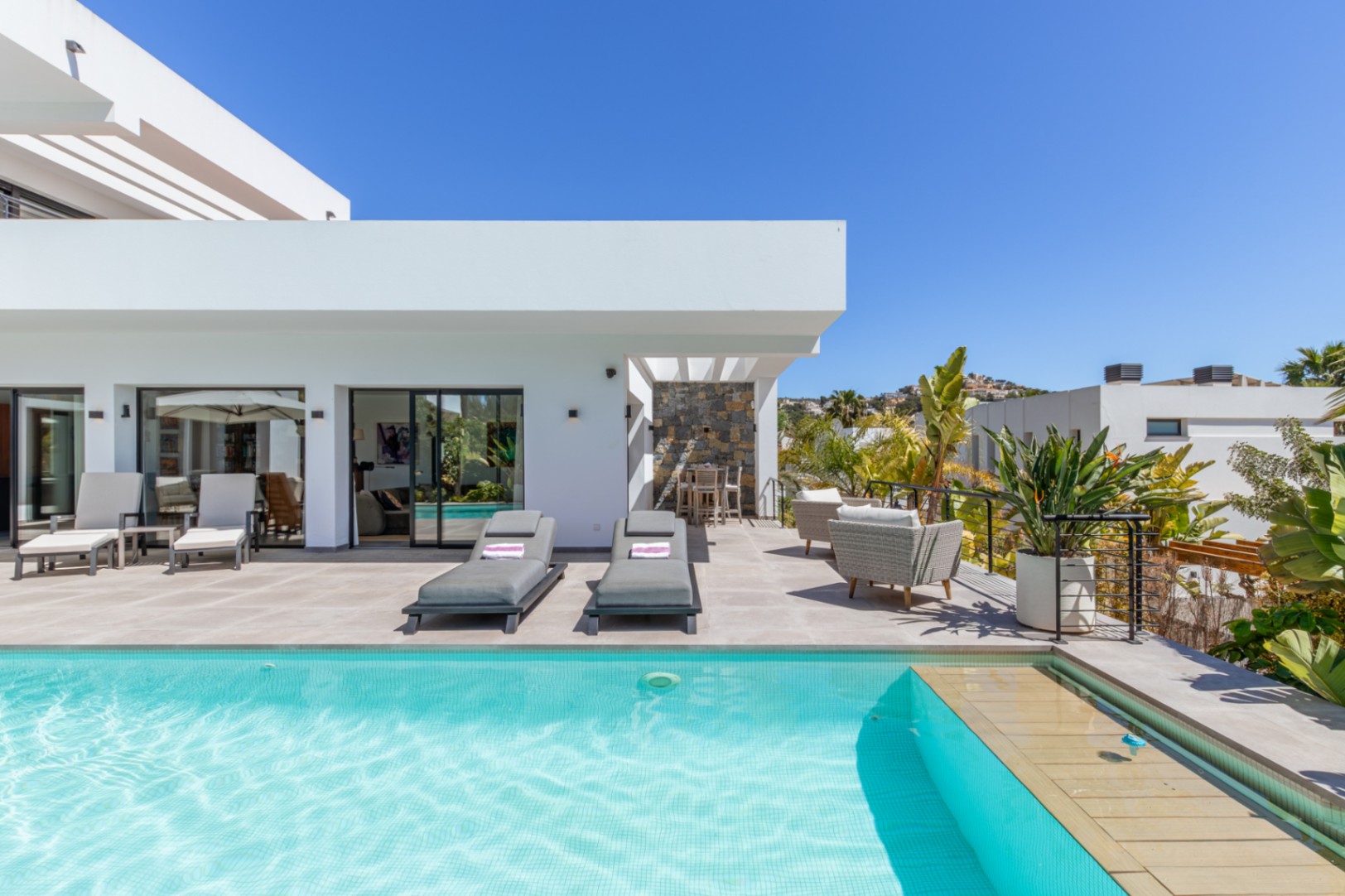 Großzügige Terrasse mit türkisfarbenem Pool, Sonnenliegen und einem Essbereich unter der Pergola, umgeben von moderner weißer Architektur und mediterraner Bepflanzung.
