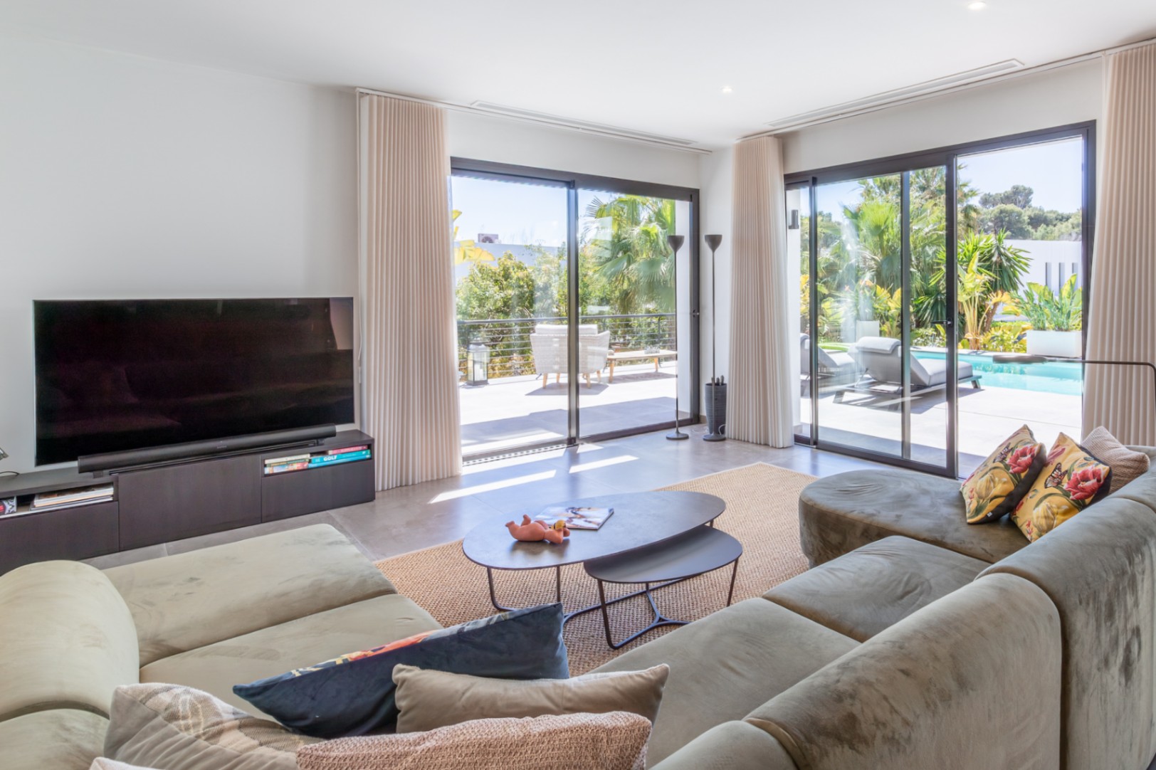 Helles Wohnzimmer mit großem Ecksofa und bodentiefen Glasschiebetüren, die direkten Zugang zur Poolterrasse und dem gepflegten Garten bieten.