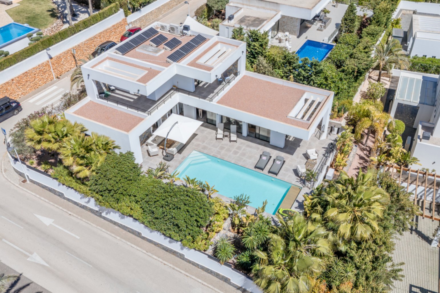 Luftaufnahme der modernen Villa mit Solarmodulen auf dem Dach, rechteckigem Pool und weitläufigen Terrassen inmitten eines eingewachsenen Gartens.