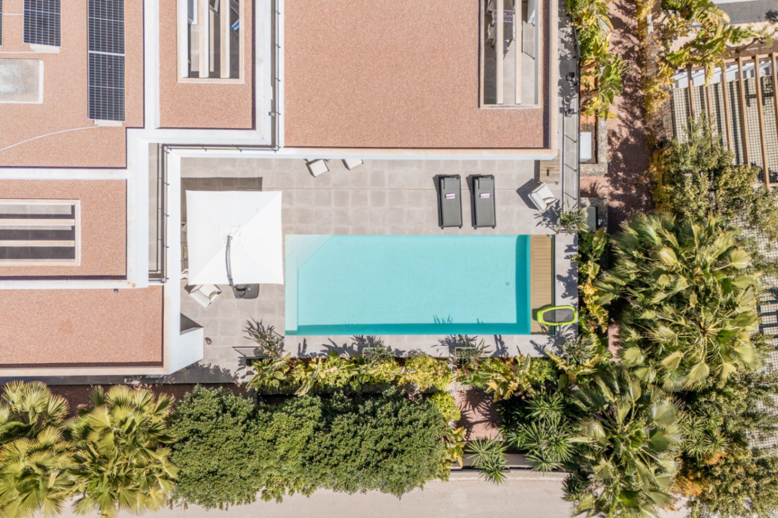 Luftaufnahme des rechteckigen Pools und der Steinterrasse, umgeben von mediterraner Bepflanzung und Solarmodulen auf dem Flachdach.