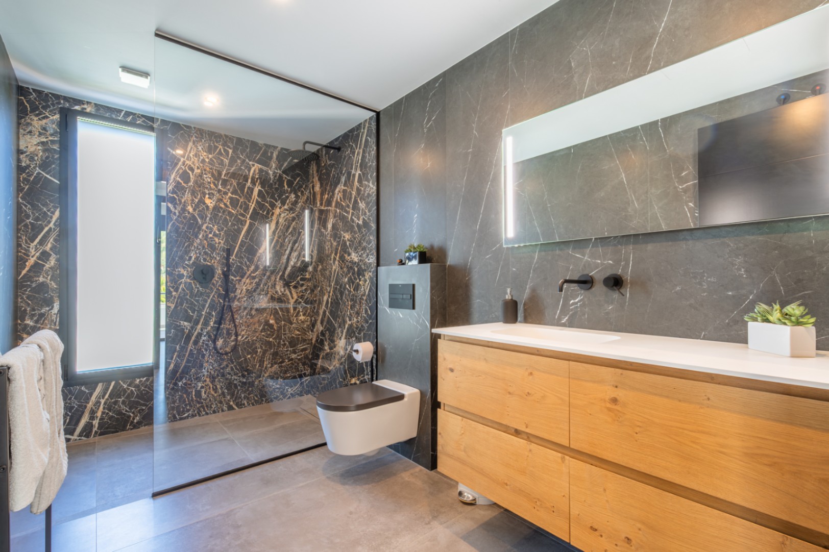 Modernes Badezimmer mit dunklen Fliesen in Marmoroptik, Waschtisch aus Naturholz und einer begehbaren Dusche mit Glaswand und schwarzen Armaturen.