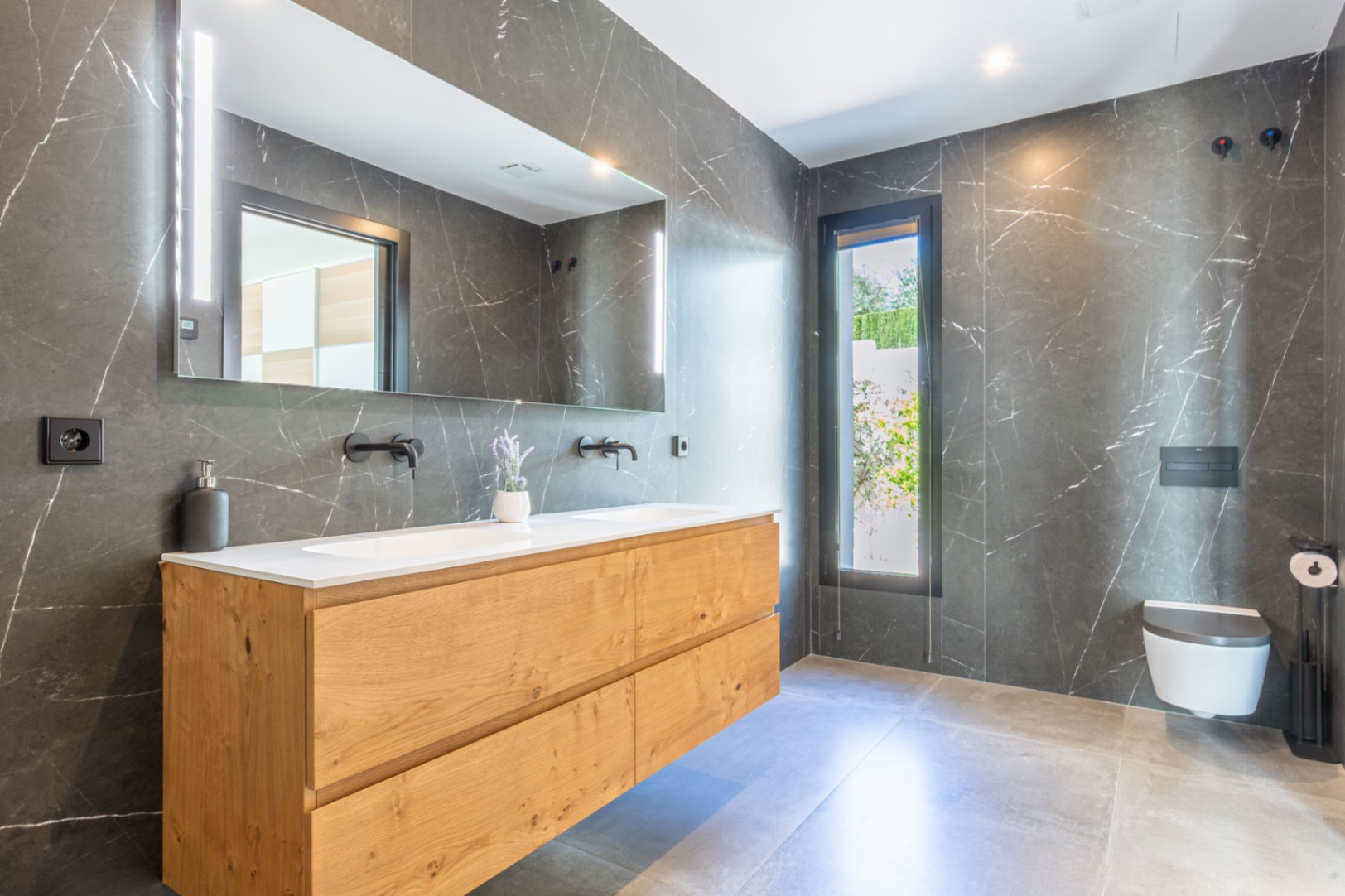Modernes Badezimmer mit dunklen Marmoreffekt-Fliesen, Doppelwaschtisch aus Holz und mattschwarzen Armaturen. Ausgestattet mit Wand-WC und großem Spiegel mit integrierter LED-Beleuchtung.