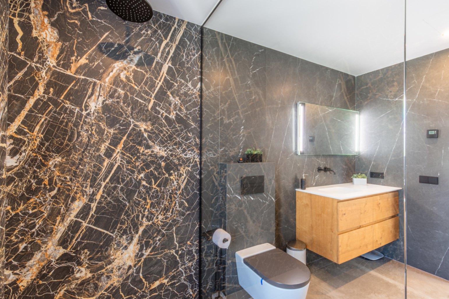 Modernes Badezimmer mit Regendusche, dunklen Marmor-Effekt-Fliesen, schwebendem Holzwaschtisch und hinterleuchtetem Spiegel.