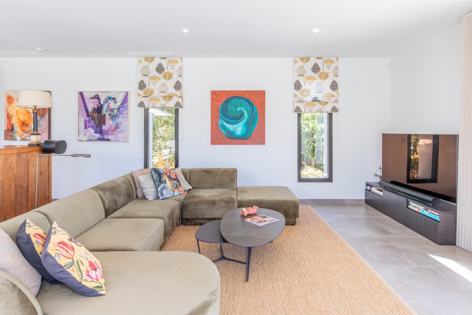 Modernes Wohnzimmer mit großem olivgrünem Ecksofa, Juteteppich und schmalen Fenstern, ergänzt durch farbenfrohe Kunstwerke an den weißen Wänden.
