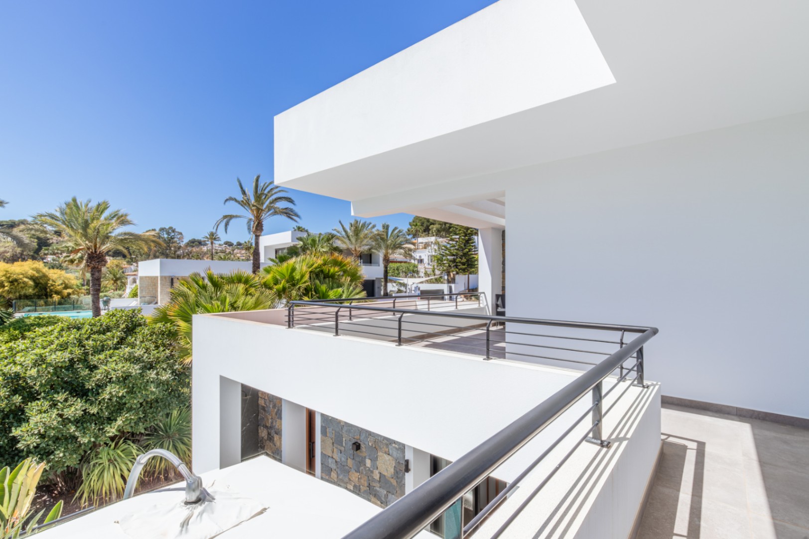 Obergeschoss-Terrasse mit schwarzem Metallgeländer und Blick auf den mediterranen Garten. Die moderne weiße Fassade wird durch Natursteinakzente ergänzt.