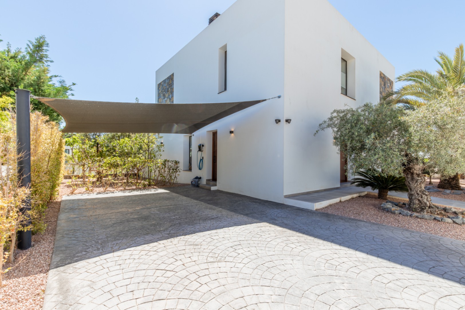 Parkplatz mit geprägtem Beton, Sonnensegel und Ladestation für Elektrofahrzeuge neben dem Seiteneingang der modernen Villa.