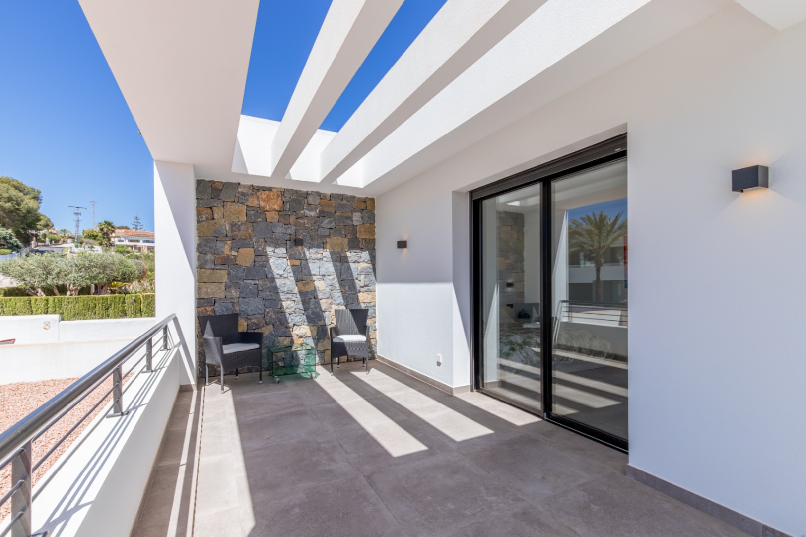 Private Terrasse mit weißen Pergola-Balken, einer Natursteinwand und großen Glasschiebetüren, die für viel Tageslicht und fließende Übergänge sorgen.