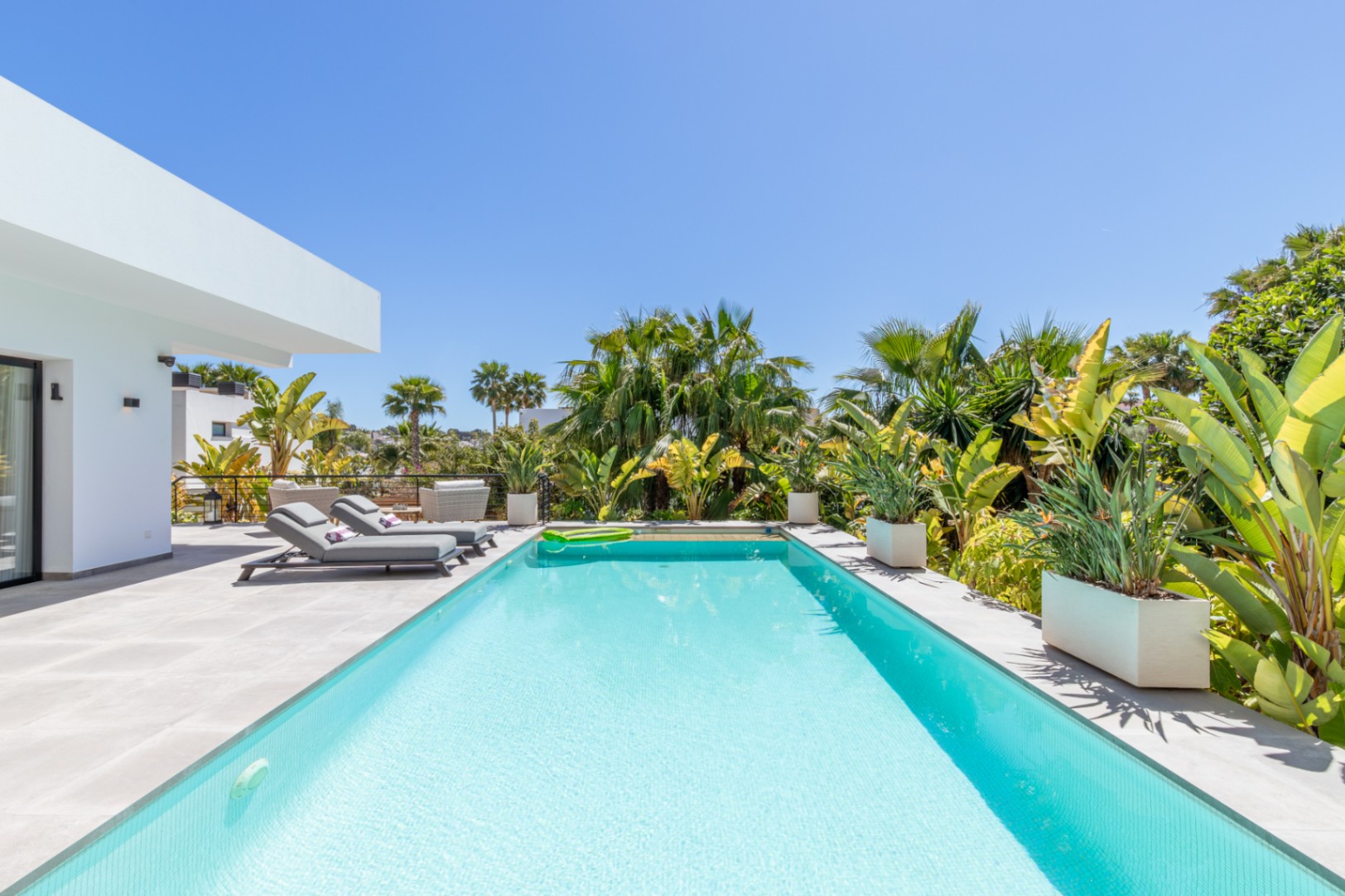 Rechteckiger Swimmingpool mit grauer Feinsteinzeug-Terrasse, umgeben von üppiger mediterraner Bepflanzung und modernen Sonnenliegen in privater Atmosphäre.