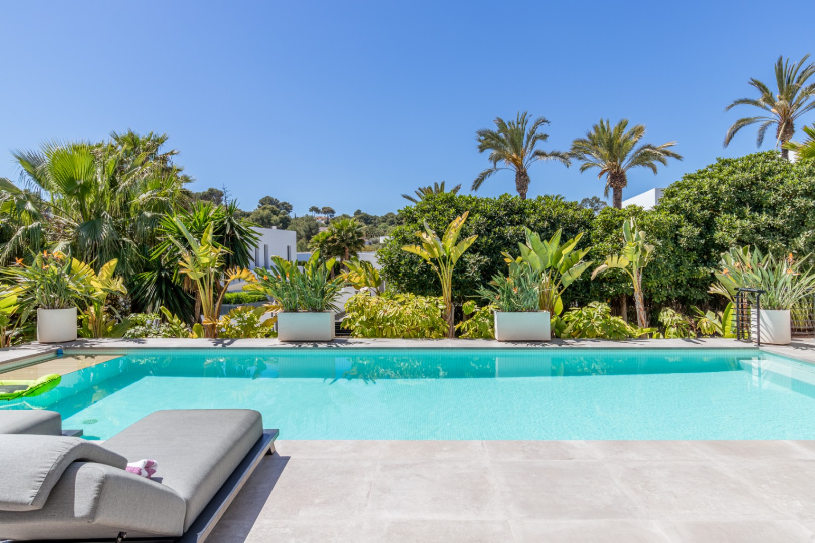Rechteckiger Swimmingpool mit heller Steinterrasse, umgeben von üppiger mediterraner Bepflanzung und Palmen, die für absolute Privatsphäre sorgen.