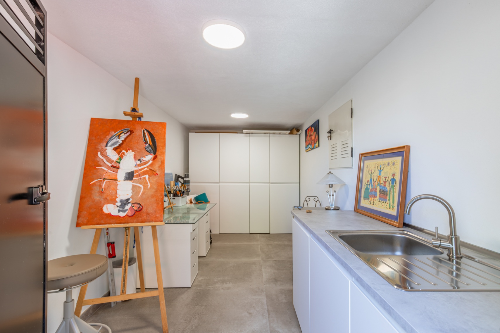 Vielseitiger Atelier- oder Hauswirtschaftsraum mit weißen Einbauschränken, Edelstahlspüle und Arbeitsbereich auf grauen Feinsteinzeugfliesen.