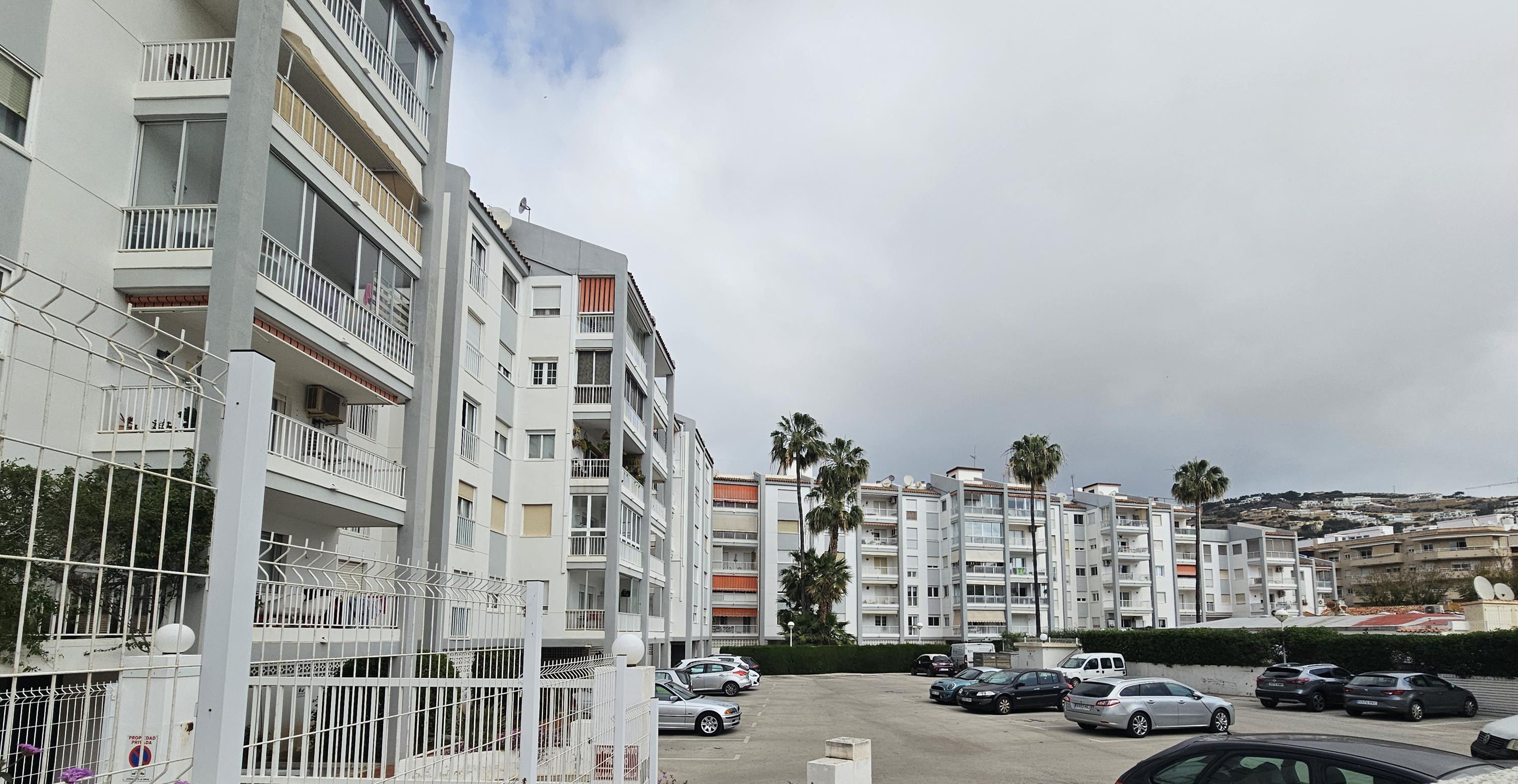 Außenansicht der Wohnanlage mit privaten Balkonen, einem eingezäunten Gemeinschaftsparkplatz und Palmen in mediterraner Umgebung.