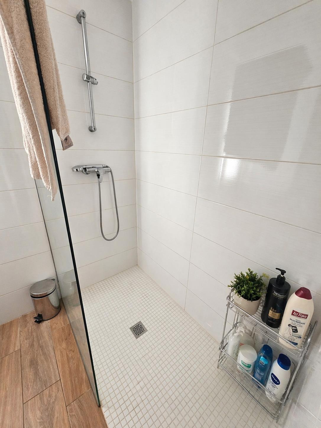Begehbare Dusche mit weißem Mosaikboden, Glastrennwand und verchromten Armaturen vor hellen, horizontalen Wandfliesen.