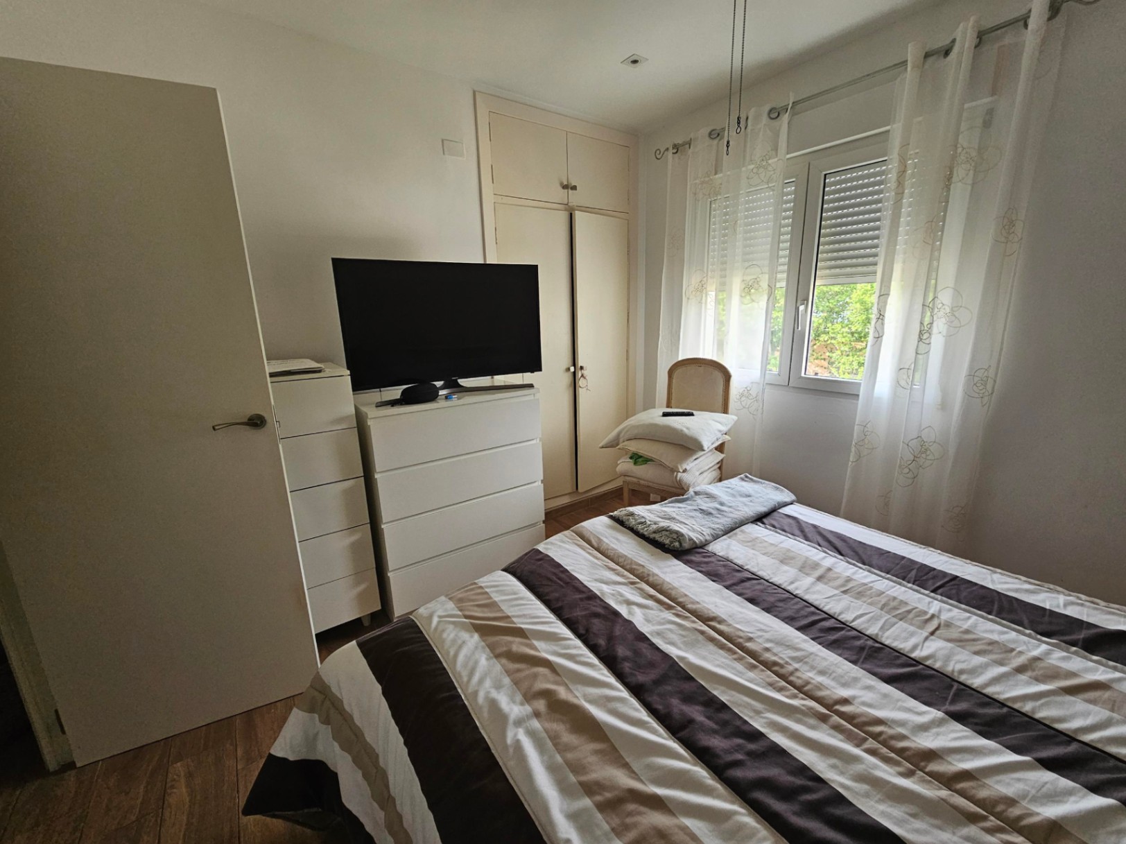 Helles Schlafzimmer mit Doppelbett, Einbauschränken, weißer Kommode mit Fernseher und Fenster mit Außenrollläden.