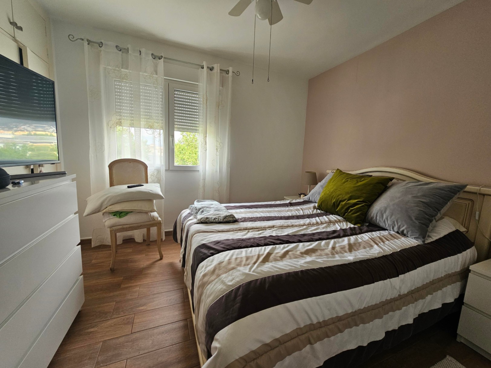 Helles Schlafzimmer mit rosa Akzentwand, Bodenfliesen in Holzoptik, Deckenventilator und Fenster mit Rollläden und transparenten Vorhängen.