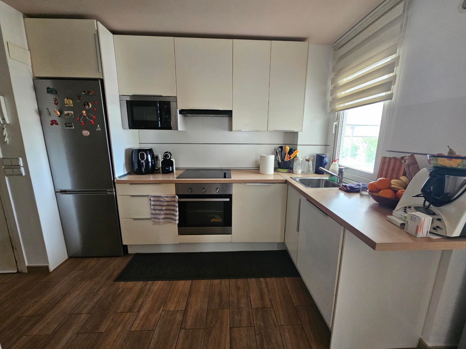 Moderne L-Küche mit weißen Schränken, Arbeitsplatte in Holzoptik, integrierten Edelstahlelektrogeräten und Boden in Holzdesign.