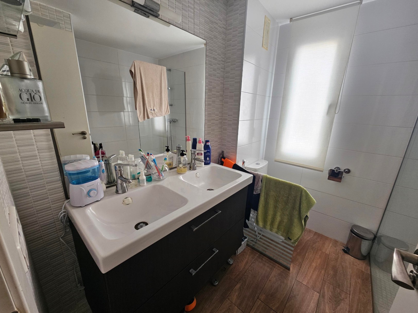Modernes Badezimmer mit Doppelwaschtisch in dunkler Optik, Feinsteinzeugboden in Holzoptik und neutralen Wandfliesen mit natürlichem Lichteinfall.