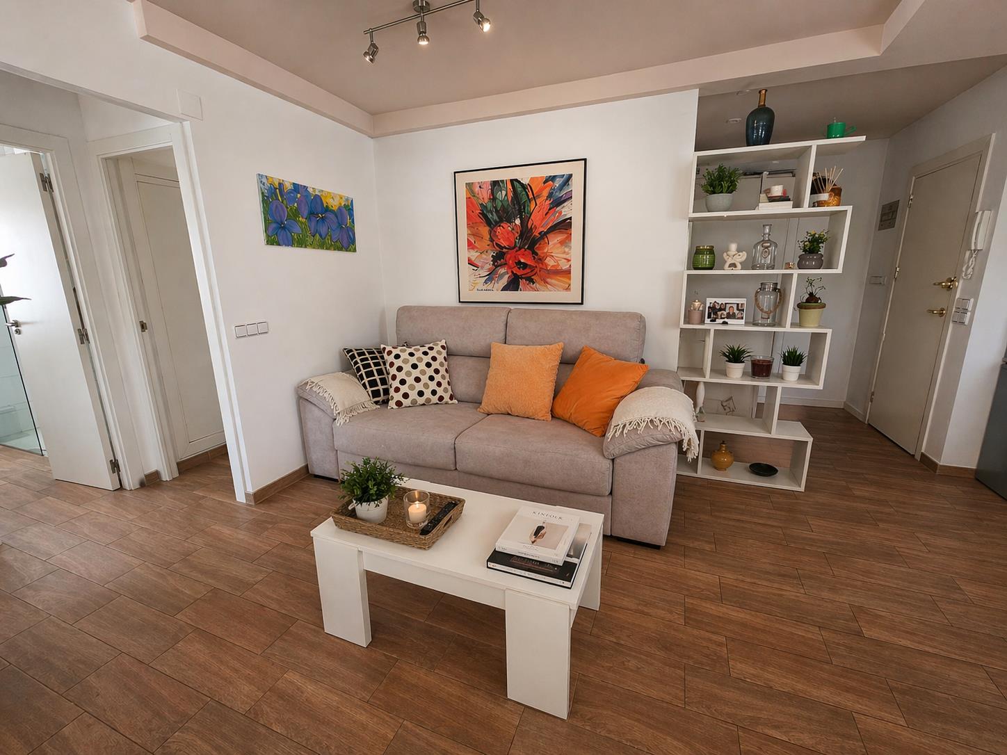 Wohnbereich mit Bodenfliesen in Holzoptik, grauem Sofa und einem modernen geometrischen Regal. Farbenfrohe Kunstwerke und Schienensystem-Beleuchtung setzen Akzente.