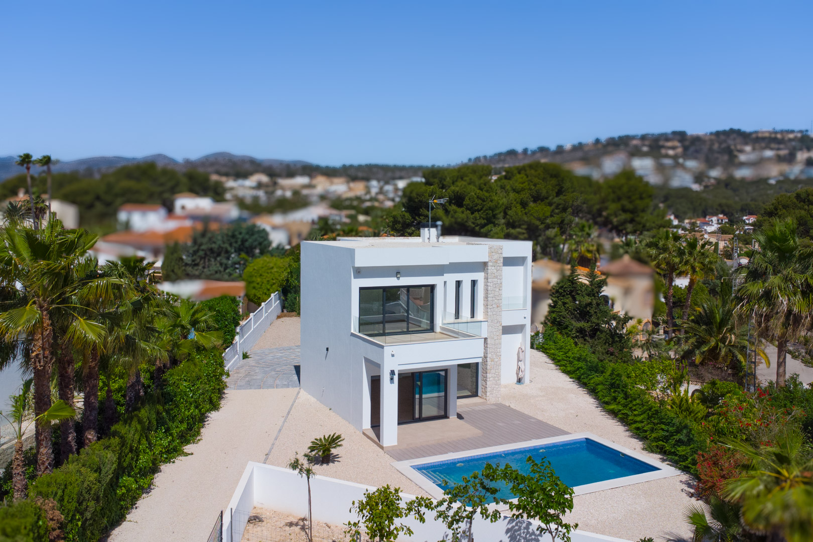 Außenansicht der modernen Villa mit weißer Fassade, Natursteinakzenten, rechteckigem Pool und Terrassen mit Glasgeländern auf einem weitläufigen, flachen Grundstück.