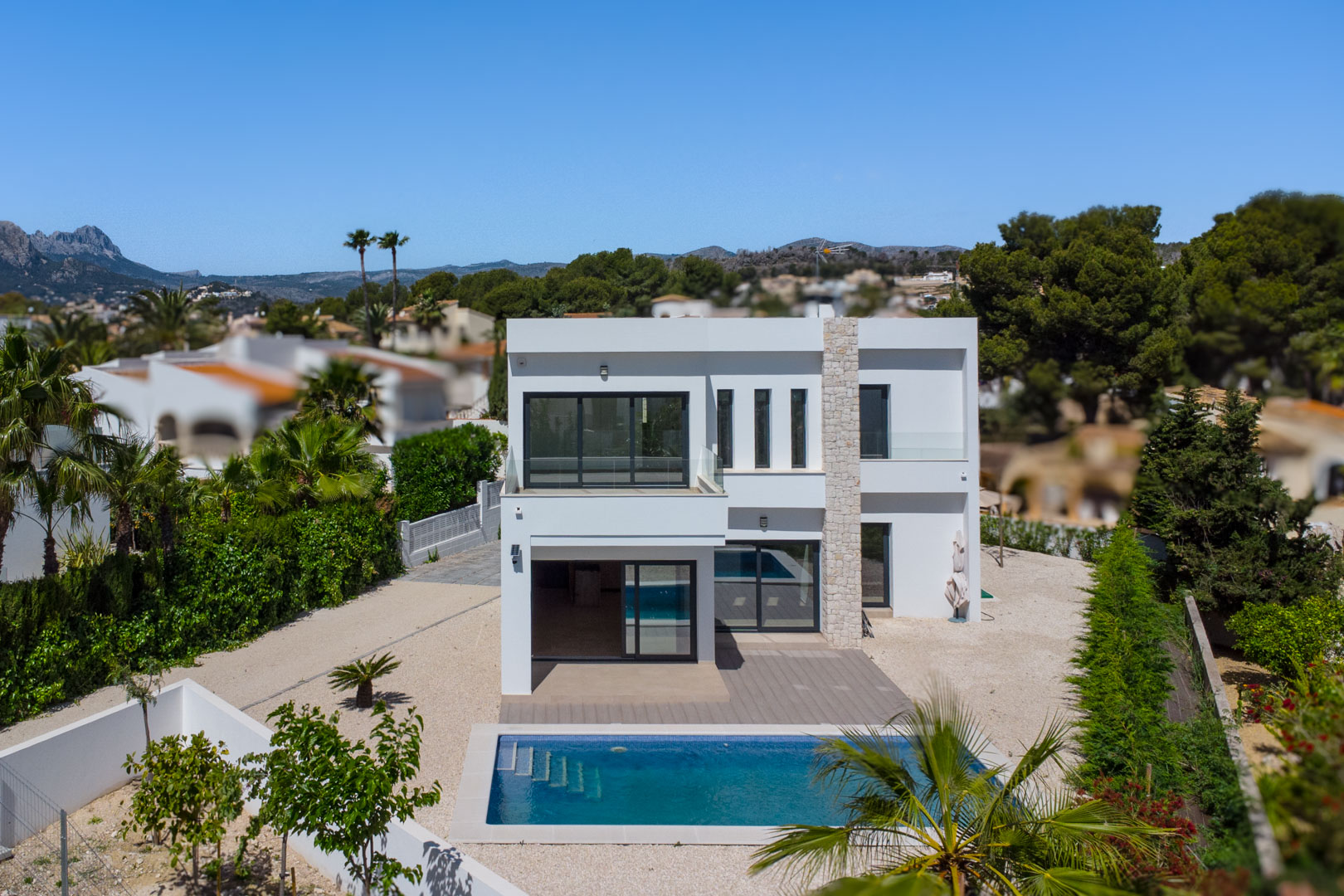 Außenansicht der modernen weißen Villa mit Natursteinpfeiler, rechteckigem Pool und großzügigen Terrassen mit Glasgeländern.