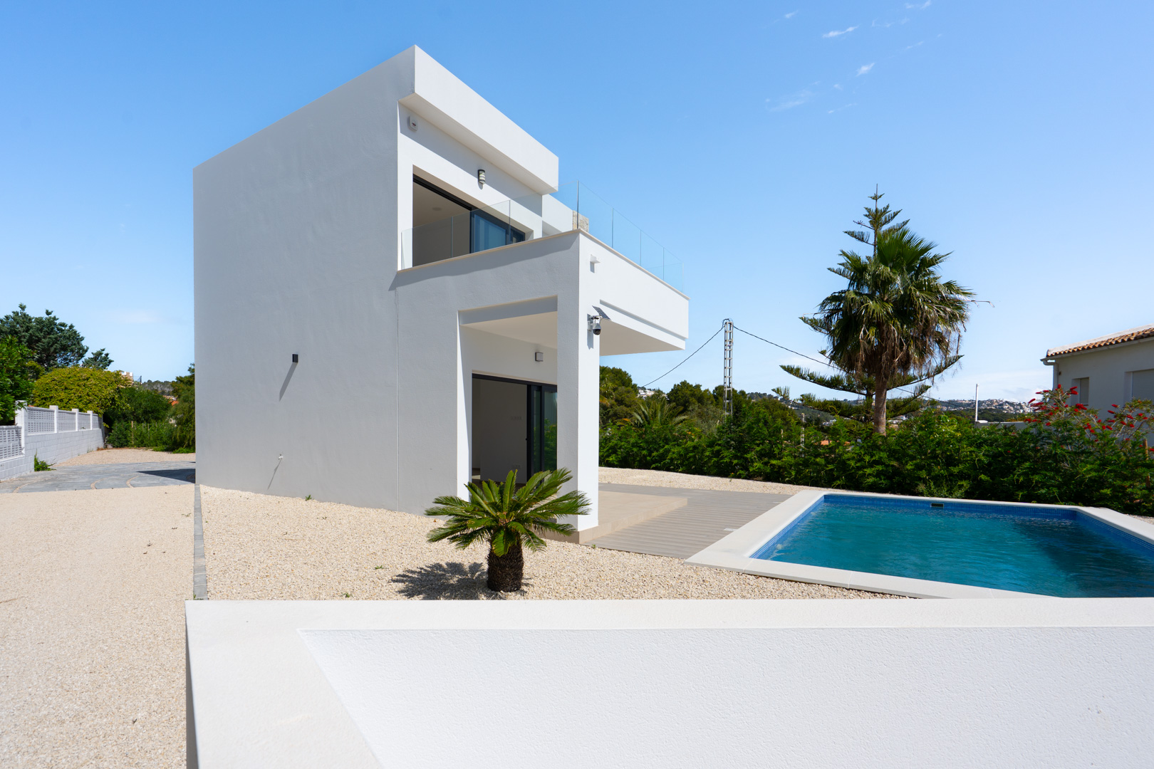 Außenansicht der modernen weißen Villa mit rechteckigem Pool, minimalistischem Kiesgarten und großzügigen Terrassen mit Glasgeländern.