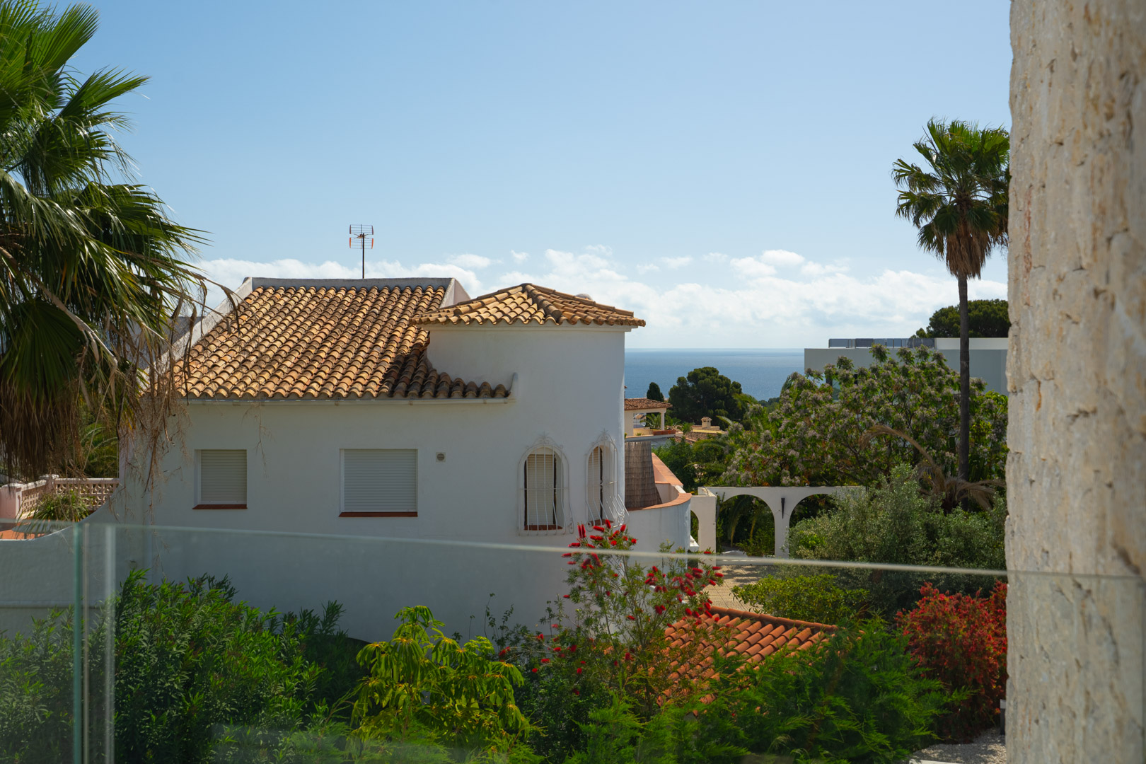 Blick auf das Mittelmeer von der oberen Terrasse mit Glasgeländer, Natursteinmauerwerk und üppiger mediterraner Bepflanzung.