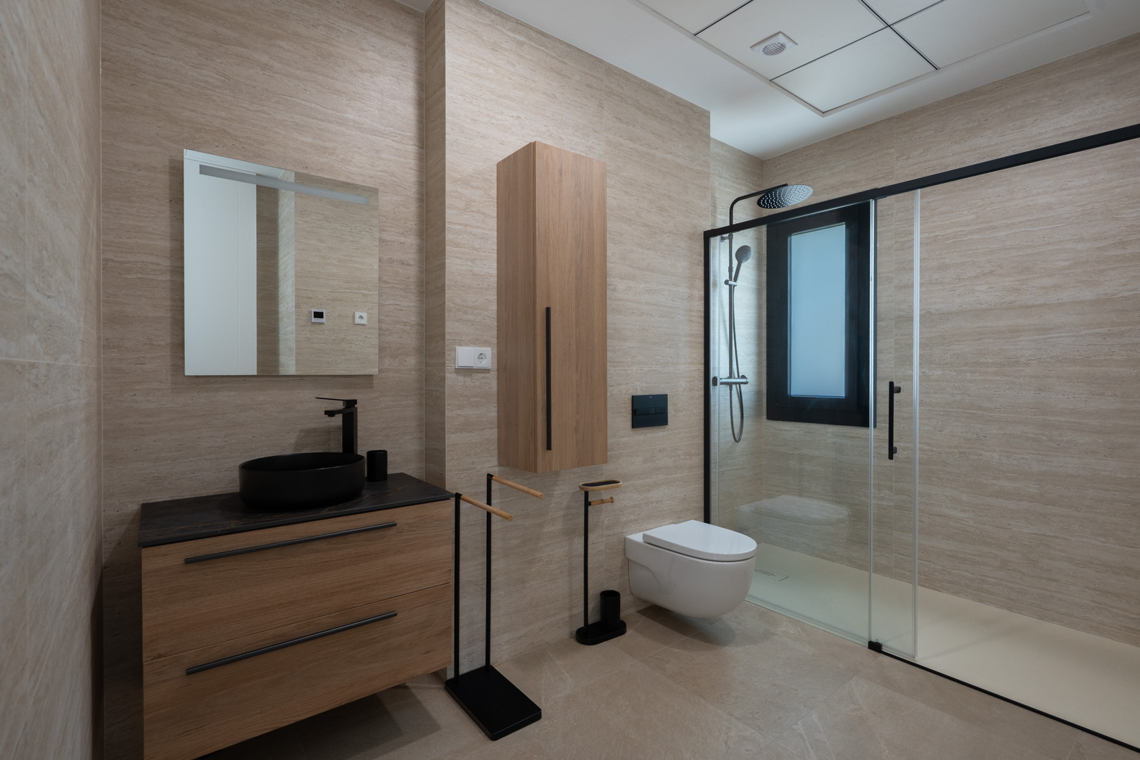 Design-Badezimmer mit Feinsteinzeug in Travertin-Optik, Holz-Waschtisch mit schwarzem Aufsatzbecken und ebenerdiger Dusche mit schwarzen Profilen.
