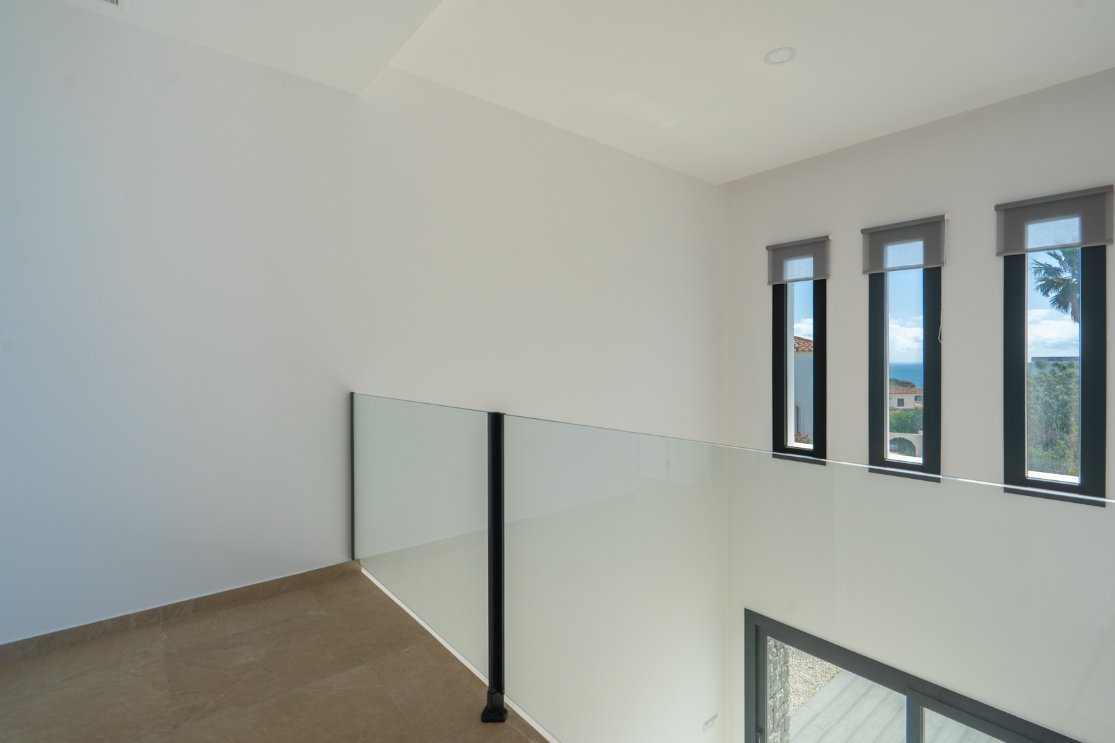 Flur im Obergeschoss mit Glasgeländer über dem Luftraum des Wohnzimmers. Drei vertikale Fenster mit Meerblick sorgen für Helligkeit und ein modernes Ambiente.