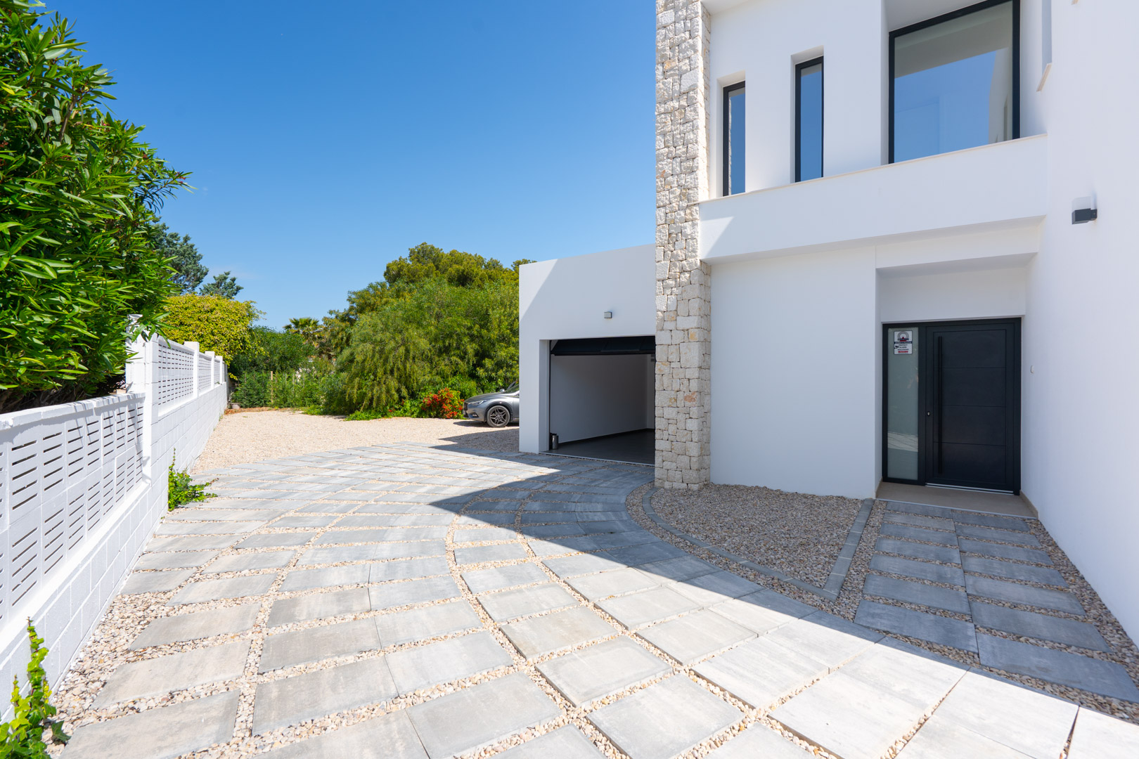 Haupteingang und Garagenzufahrt mit gepflastertem Weg und Kieselementen, akzentuiert durch eine weiße Fassade und eine Natursteinwand.