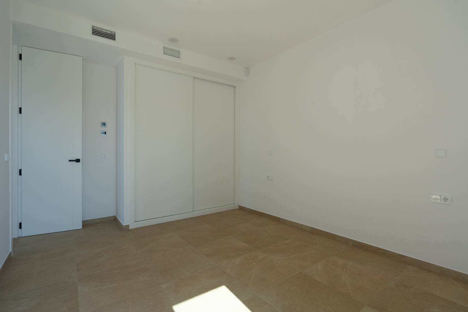 Helles Schlafzimmer mit erdfarbenen Feinsteinzeugfliesen, Einbauschrank mit weißen Schiebetüren und integrierter Kanalklimaanlage.