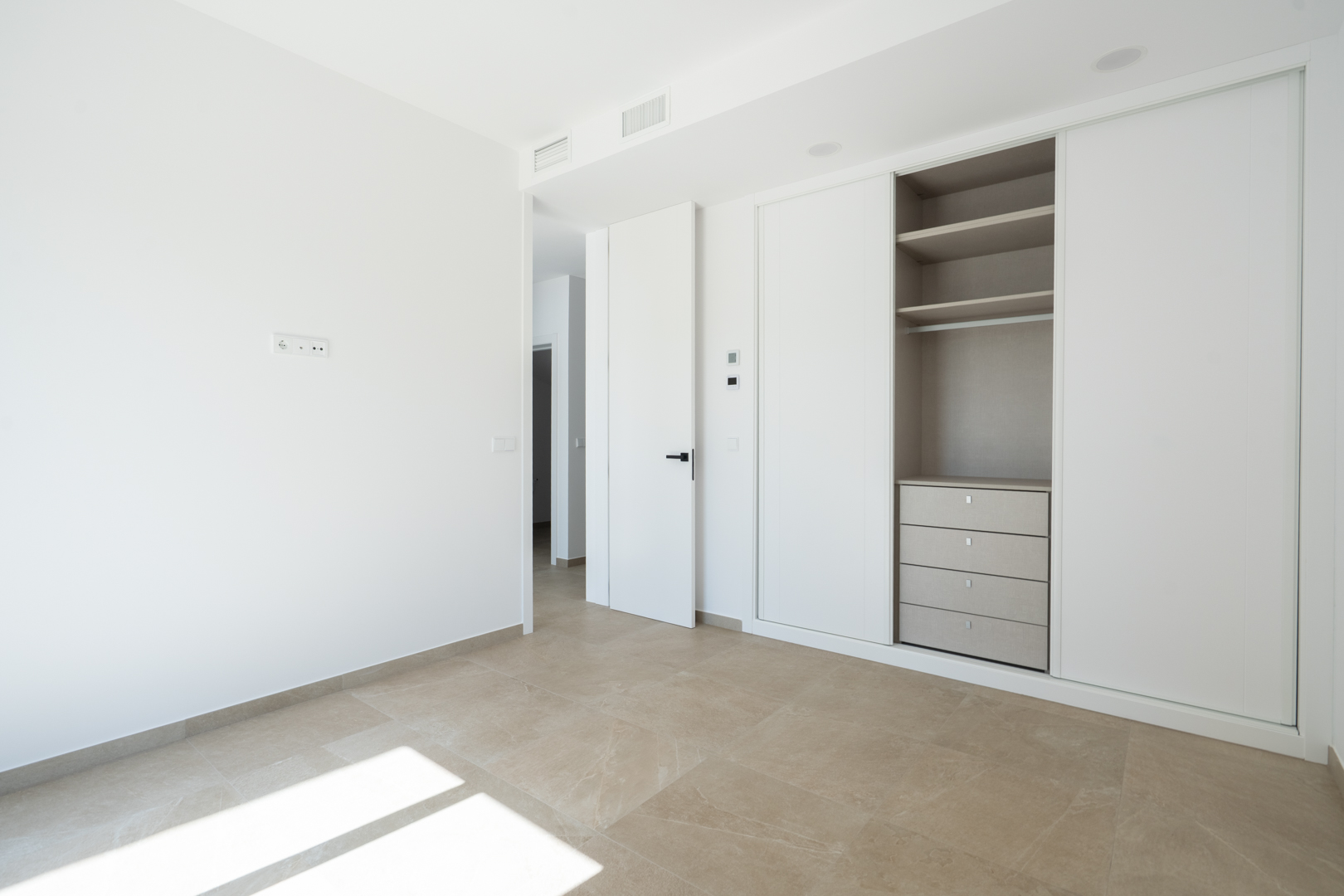 Helles Schlafzimmer mit weißen Einbauschränken, Schiebetüren, strukturierter Inneneinteilung mit Schubladen und großformatigen Feinsteinzeugfliesen.