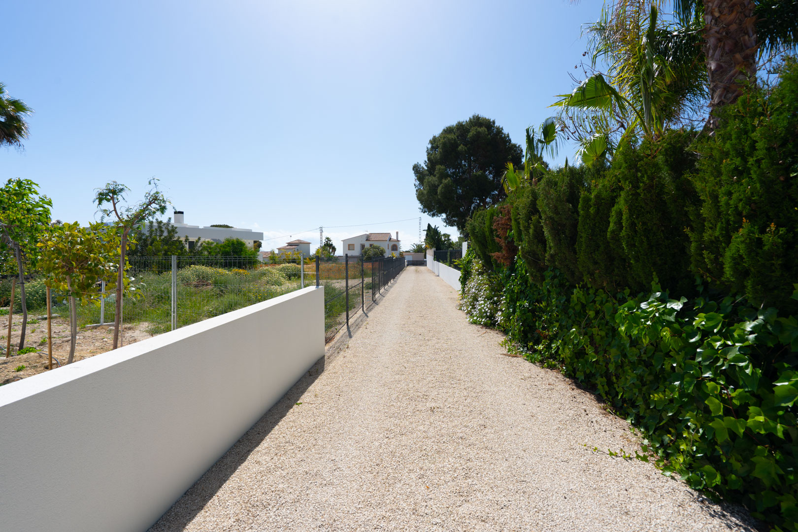 Lange Kiesauffahrt, begrenzt durch eine weiße Mauer und eine dichte Hecke, die das weitläufige, ebene Grundstück der Villa erschließt.