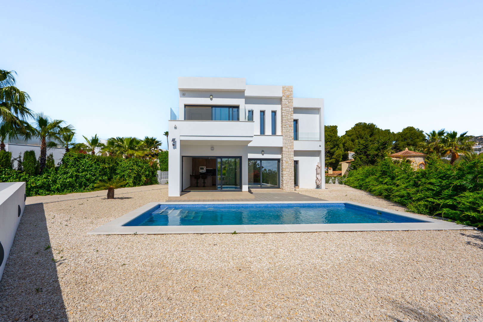 Moderne zweistöckige Villa mit weißer Fassade, Natursteinakzenten, rechteckigem Pool und Kiesgarten, umgeben von mediterraner Bepflanzung.