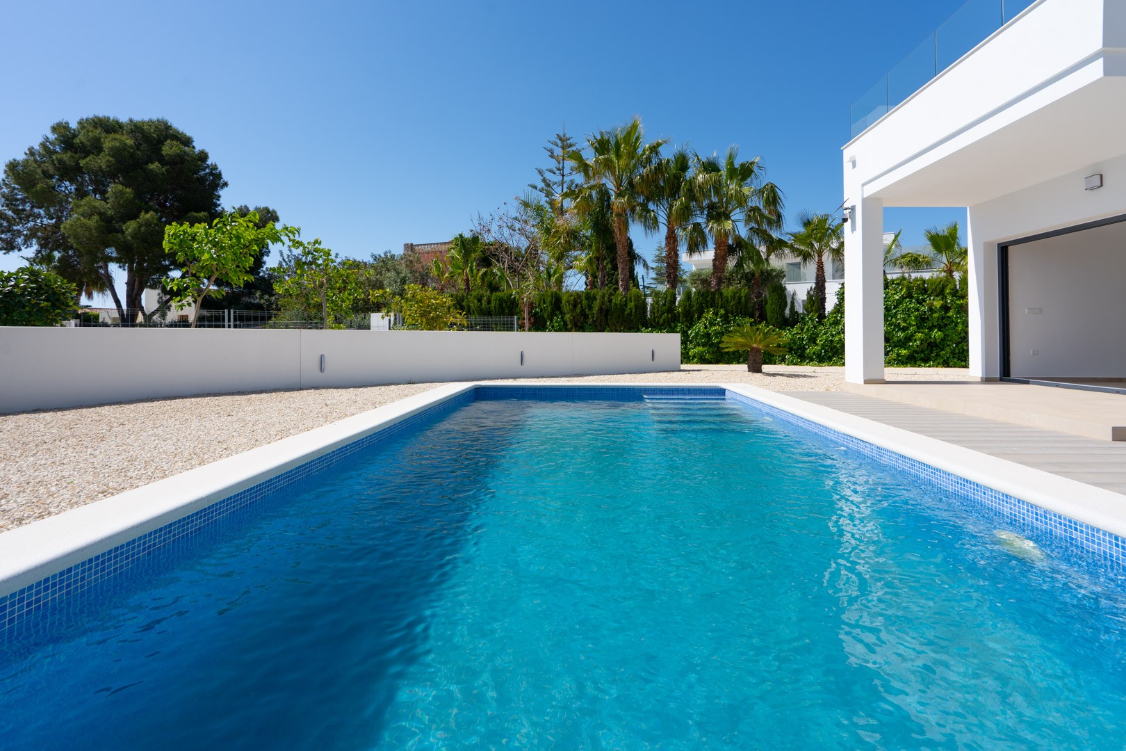 Rechteckiger Pool mit blauen Mosaikfliesen, umgeben von weißem Zierkies und modernen Mauern mit integrierter Beleuchtung vor Palmenkulisse.