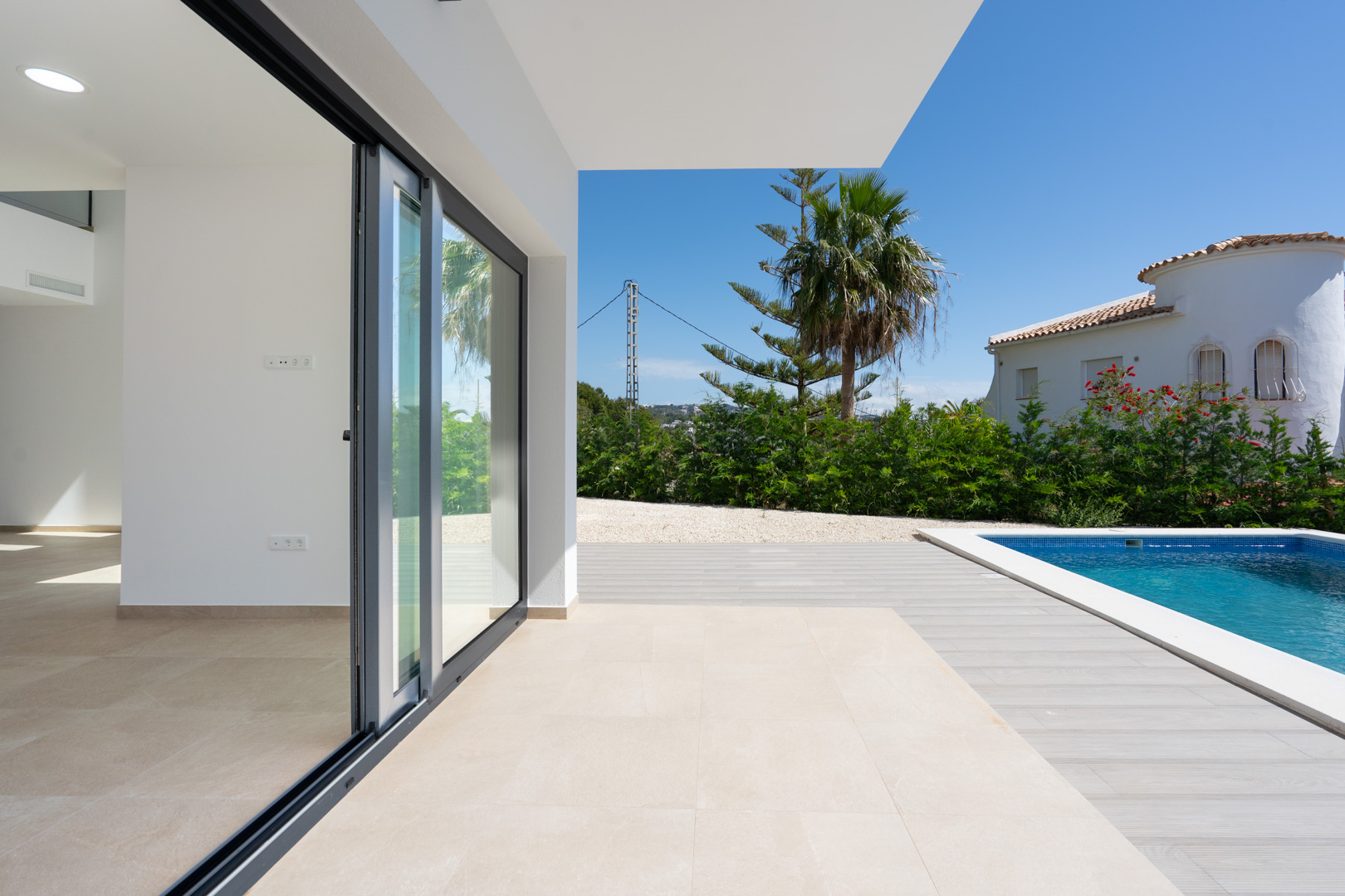 Überdachte Terrasse mit großformatigen Feinsteinzeugfliesen, die nahtlos in den Poolbereich mit Holzoptik-Deck und mediterraner Bepflanzung übergeht.