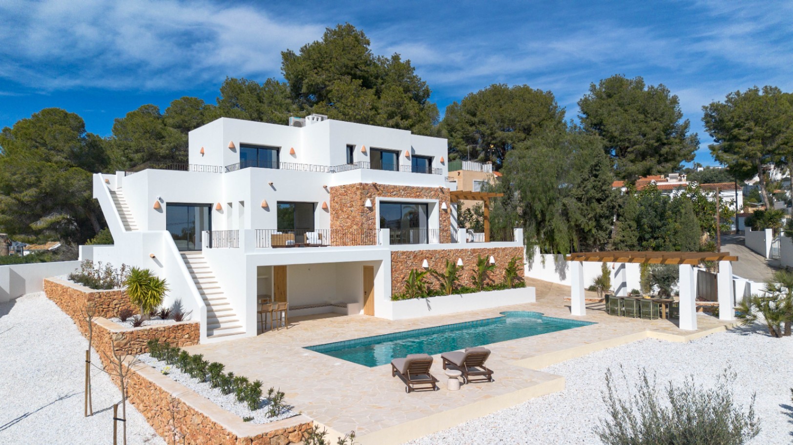 Außenansicht der modernen Villa mit weißen Fassaden, Natursteinwänden, einem rechteckigen Pool und mehreren Terrassen inmitten mediterraner Bepflanzung.