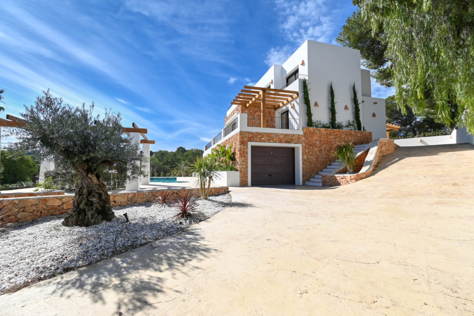 Außenansicht der Villa mit Natursteinfassade, integrierter Garage und gepflasterter Einfahrt, gesäumt von einem Olivenbaum und mediterraner Bepflanzung.