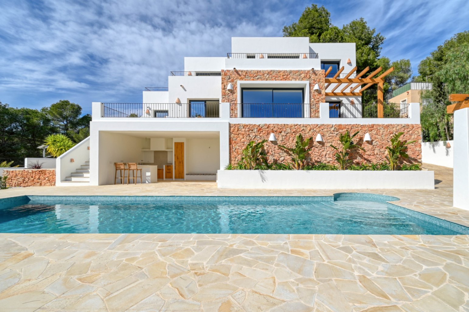 Außenansicht mit blauem Mosaikpool, Natursteinterrasse und mehrstöckiger Fassade, die Trockensteinmauern, eine Außenküche und eine Holzpergola harmonisch kombiniert.