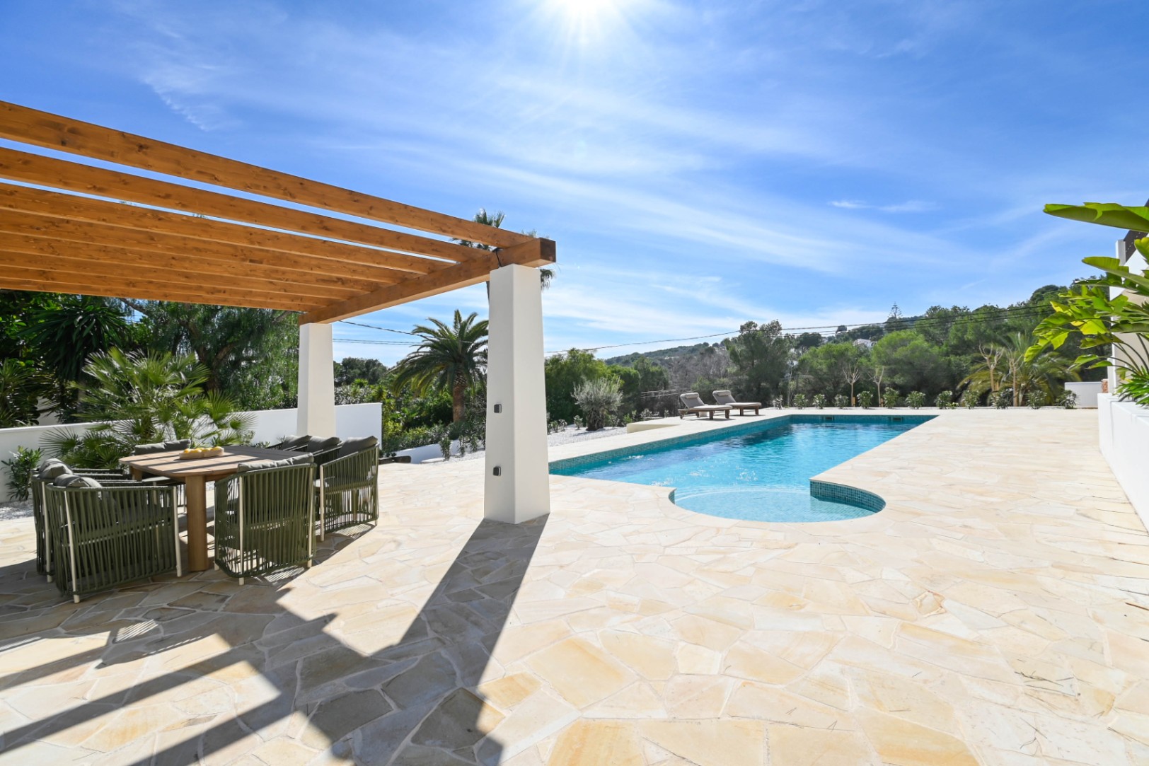 Außenbereich mit Natursteinpflaster, einer Holzpergola mit Essbereich und einem 13 Meter langen Pool, umgeben von mediterraner Bepflanzung.