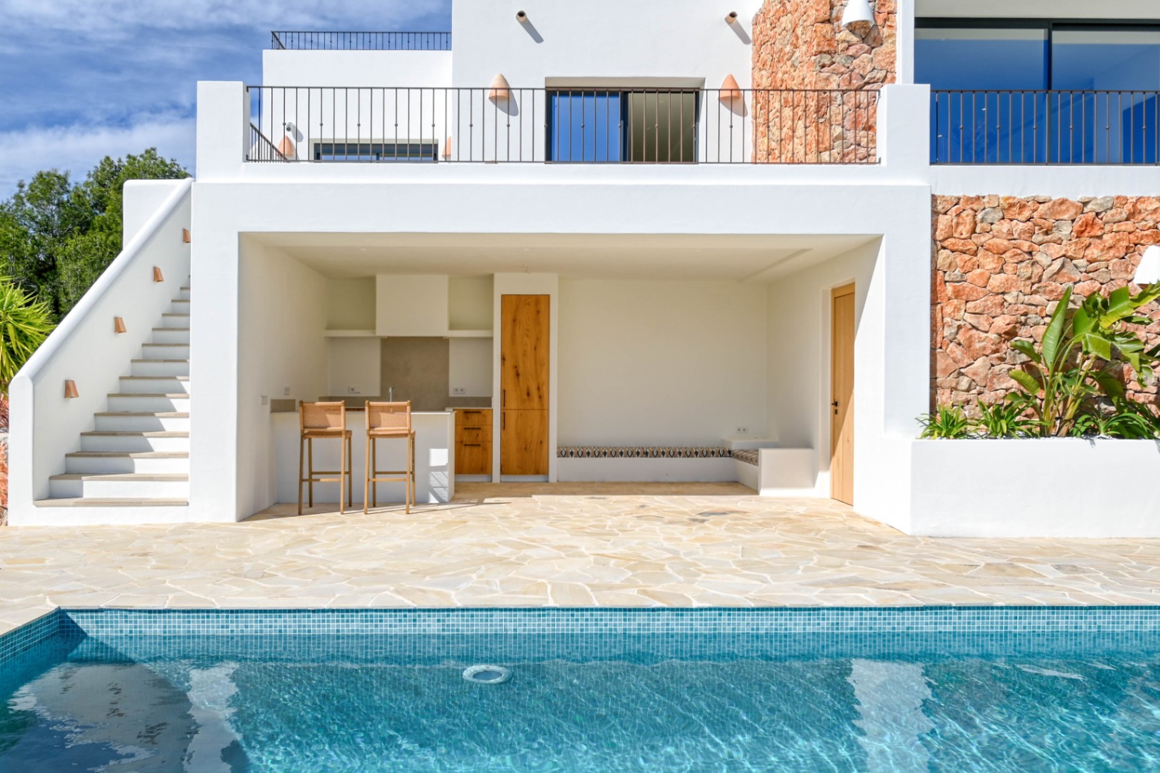 Außenküche und Loungebereich am Pool mit Natursteinwänden, Holzakzenten und einer Terrasse aus Natursteinplatten unter einer modernen weißen Fassade.