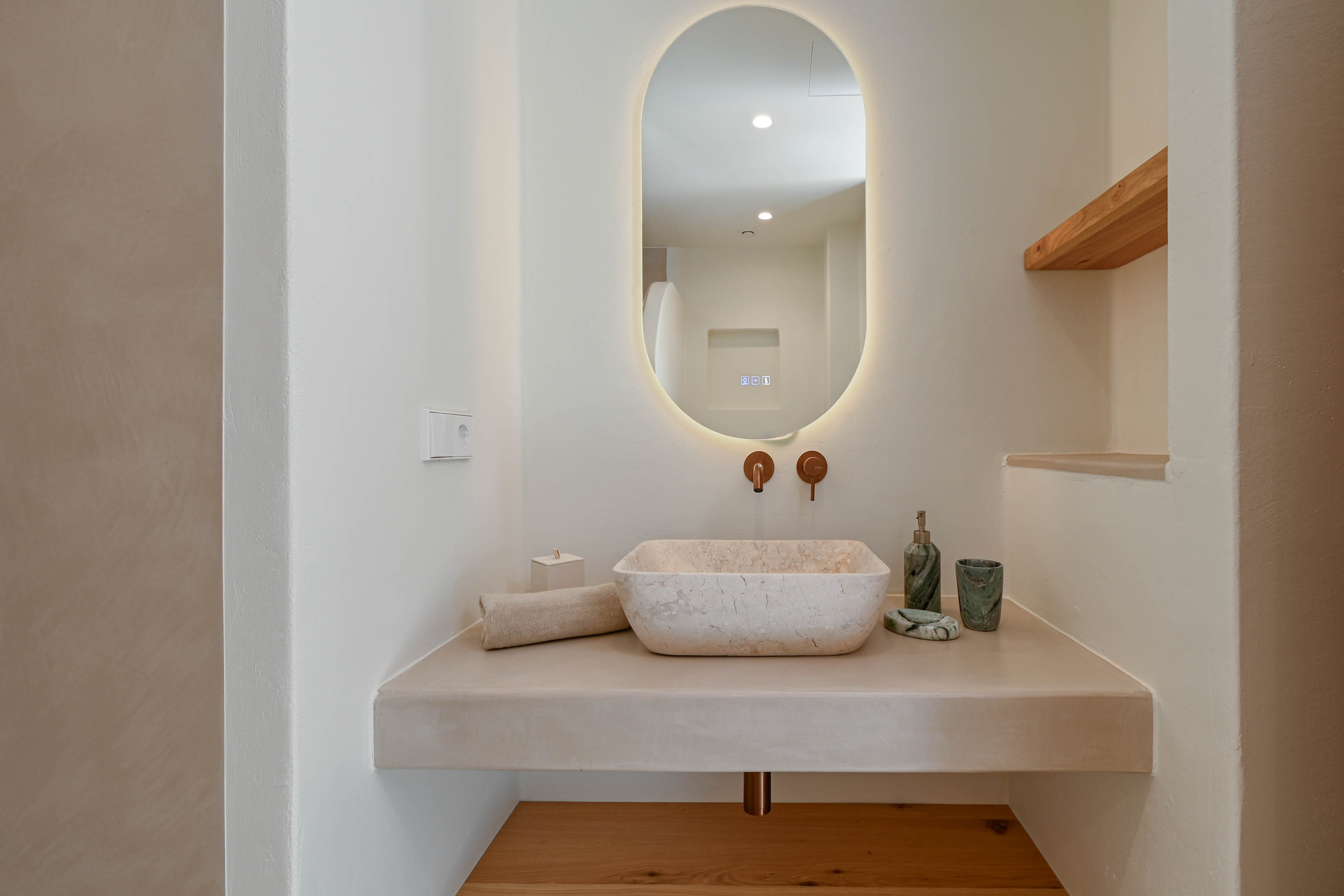 Designer-Badezimmer mit Naturstein-Aufsatzwaschbecken auf Mikrozement-Waschtisch, Unterputzarmaturen in Kupfer und hinterleuchtetem Ovalspiegel.