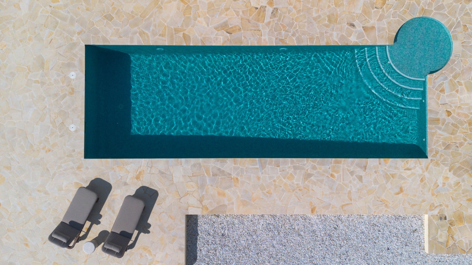 Draufsicht auf den 13 Meter langen Pool mit türkisfarbenen Mosaikfliesen, integrierter Rundtreppe und einer Terrasse aus unregelmäßigen Natursteinplatten.