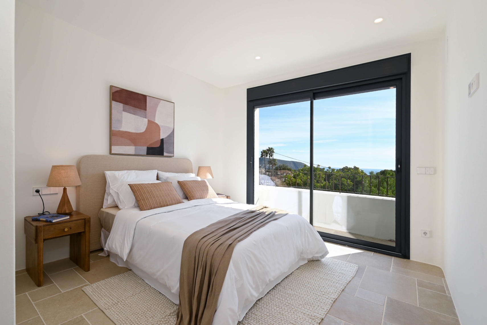 Helles Schlafzimmer mit Natursteinboden und großen Glasschiebetüren, die auf eine private Terrasse mit weitem Blick auf die mediterrane Landschaft führen.